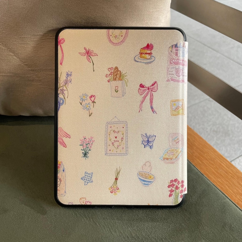 May include: A cream-coloured e-reader case with a black border, featuring a repeating pattern of watercolour illustrations. The design includes flowers, bows, a cupcake, and other whimsical elements. The case is open, revealing the screen.
