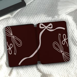 Könnte beinhalten: Eine dunkel-kastanienbraune E-Reader-Hülle mit einem hellrosa Linienmuster aus mehreren dekorativen Schleifen und Bändern. Die Hülle ist in geöffneter Position dargestellt und zeigt das Innendesign, das die Ästhetik der Außenseite widerspiegelt.