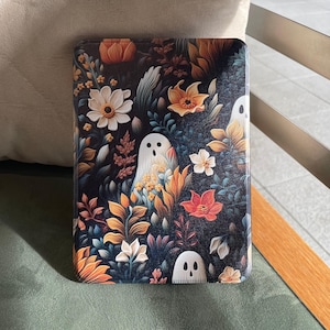 May include: A rectangular e-reader case featuring a dark background with a dense pattern of colorful flowers, foliage, and multiple white ghost figures with black eyes and mouths. The design includes orange, yellow, and red floral elements.