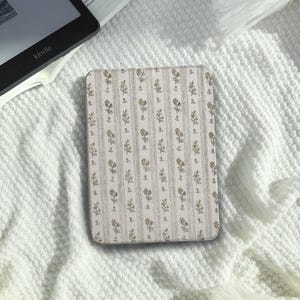 Puede incluir: Una funda rectangular para lector electrónico con fondo beige claro, rayas verticales y un patrón repetitivo de pequeñas ramas florales en tonos verde y marrón apagados. La funda tiene esquinas redondeadas y una superficie lisa y plana.