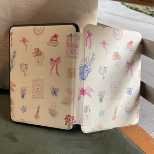 May include: A black tablet case with a cream-coloured cover. The cover has a repeating pattern of watercolour illustrations, including bows, flowers, cakes, and framed pictures. The case is open, revealing the interior design.