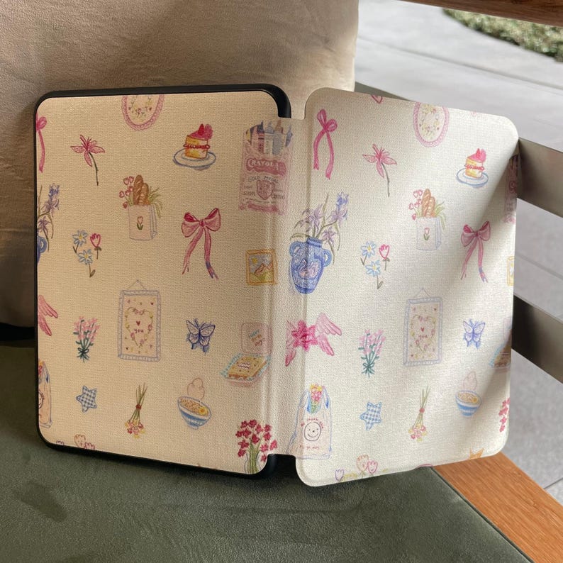 May include: A black tablet case with a cream-coloured cover. The cover has a repeating pattern of watercolour illustrations, including bows, flowers, cakes, and framed pictures. The case is open, revealing the interior design.