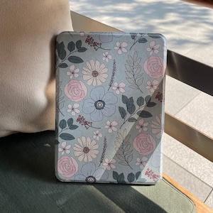 Può includere: Una custodia per tablet con motivo floreale. La custodia ha uno sfondo azzurro chiaro adornato con un motivo di fiori rosa e bianchi, insieme a foglie e steli verdi. La custodia rettangolare è probabilmente realizzata in un materiale liscio.
