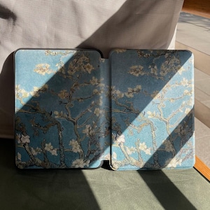 May include: An open e-reader case with a blue cover, featuring a floral design inspired by Van Gogh's "Almond Blossoms". The case has a black border and a textured finish. The design shows white blossoms and branches against a blue backdrop.