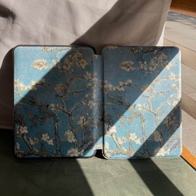 May include: An open e-reader case with a blue cover, featuring a floral design inspired by Van Gogh's "Almond Blossoms". The case has a black border and a textured finish. The design shows white blossoms and branches against a blue backdrop.