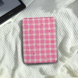 May include: A pink and white gingham pattern Kindle cover with small red star accents. The cover is rectangular and appears to be made of a soft material. The cover is resting on a white, textured surface.
