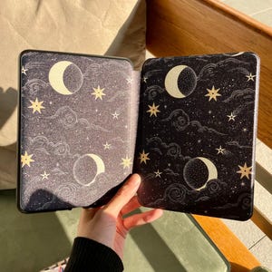 May include: An open black e-reader case with a celestial design. The case features a night sky pattern with crescent moons, stars, and clouds in gold and white. The case is held open, revealing the interior design.