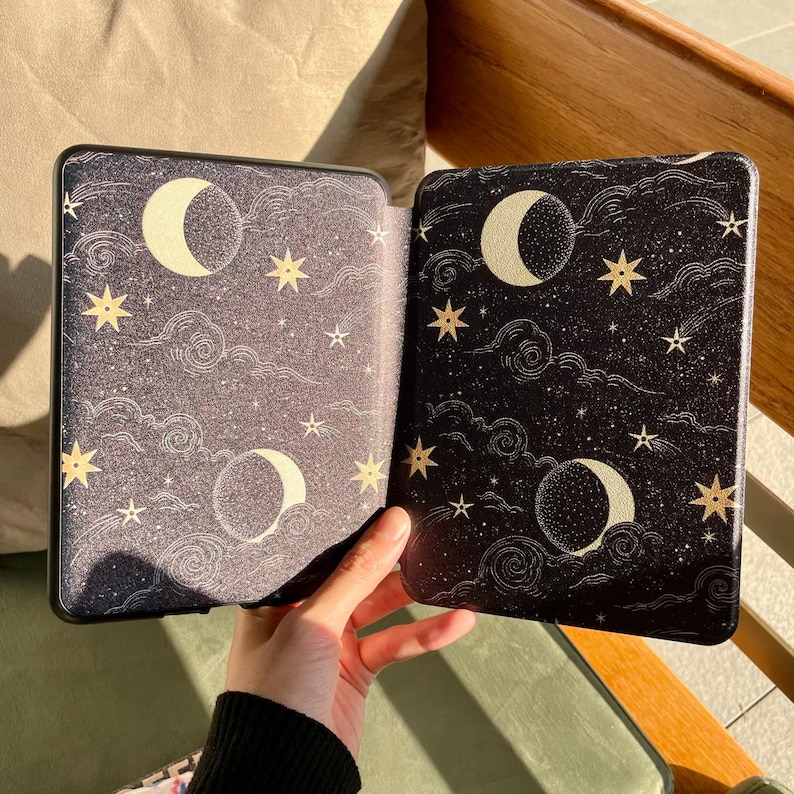 May include: An open black e-reader case with a celestial design. The case features a night sky pattern with crescent moons, stars, and clouds in gold and white. The case is held open, revealing the interior design.