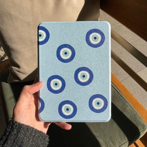Könnte beinhalten: Eine rechteckige E-Reader-Hülle mit hellblauem, strukturiertem Hintergrund und einem sich wiederholenden Muster aus dunkelblauen, hellblauen und weißen kreisförmigen Nazar-Amulett-Motiven, die über die gesamte Vorderseite verteilt sind.