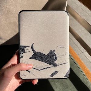 Puede incluir: Una funda de tableta ilustrada en blanco y negro con un gato negro tumbado en un libro. La funda tiene un fondo beige claro y un borde negro. Tambi&eacute;n se ilustran un bol&iacute;grafo, un tintero y una taza de caf&eacute;.