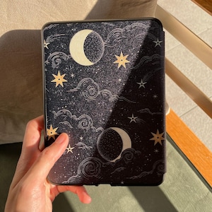 May include: A black e-reader case with a celestial design. It features a crescent moon, stars, and clouds. The case has a dark background with white dots and gold stars. The rectangular case has a black border. The design evokes a night sky.