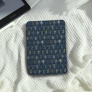Può includere: Una custodia rettangolare per e-reader con sfondo blu scuro e un motivo a reticolo dorato ripetuto. All'interno di ogni sezione del reticolo si trovano piccole illustrazioni dettagliate di vari fiori e fogliame nei toni del blu, bianco e giallo.