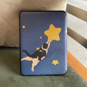 Könnte beinhalten: Tablethülle mit einer schwarz-weißen Katze, die nach einem großen gelben Stern auf blauem Hintergrund greift. Das Design umfasst kleinere gelbe Sterne. Schwarzer Rand.