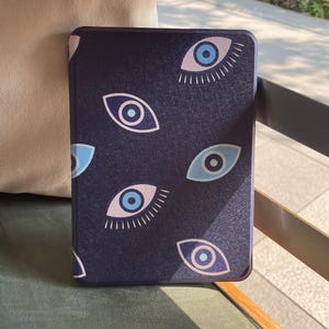 Könnte beinhalten: Eine marineblaue Tablethülle mit einem sich wiederholenden Muster aus stilisierten blauen und rosa Augen. Die rechteckige Hülle besteht aus einem strukturierten Material. Das Design ist modern und auffällig.