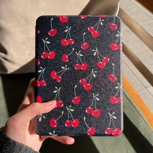 Puede incluir: Una funda negra para tableta con un patrón repetido de cerezas rojas y hojas verdes. La funda es rectangular y tiene esquinas redondeadas. Las cerezas son de color rojo vibrante y las hojas son de color verde claro.