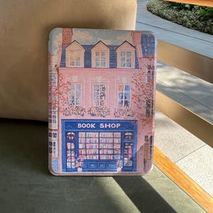 Könnte beinhalten: Eine rosa-blaue Hülle mit einer detaillierten Illustration eines Buchladens. Der Laden hat eine blaue Markise mit den Worten "BOOK SHOP" und ist von rosa Blumen und Bäumen umgeben. Die Hülle dient zum Schutz eines Tablets oder E-Readers.