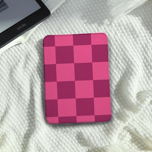 Peut inclure: Un étui Kindle à motif de damier rose et magenta. L'étui est rectangulaire et recouvre l'appareil. L'étui repose sur une couverture blanche texturée. Un appareil Kindle est partiellement visible dans le coin supérieur gauche.