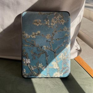 May include: A rectangular case with a print of Van Gogh's "Almond Blossoms" artwork. The case has a black border and a blue background with white and yellow flowers and branches. It is likely designed for a tablet or e-reader.