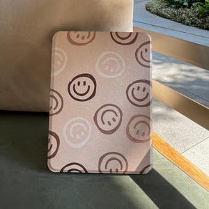 Könnte beinhalten: Rechteckige Hülle mit einem sich wiederholenden Muster aus Smiley-Gesichtern in Brauntönen auf hellbraunem Hintergrund. Die Hülle, wahrscheinlich für einen E-Reader oder ein Tablet, hat abgerundete Ecken. Das Design ist einfach und fröhlich.