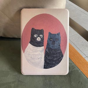 Könnte beinhalten: Eine rechteckige E-Reader-Hülle mit einer grafischen Illustration von zwei Katzen, die gestrickte Sturmhauben tragen. Eine Katze ist weiß mit schwarzer Sturmhaube, die andere schwarz mit blauer Sturmhaube, vor einem kreisförmigen altrosa Hintergrund.