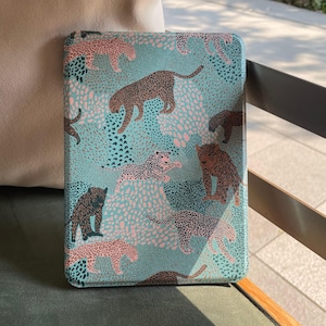 Pode incluir: Uma capa retangular turquesa com um padrão repetido de leopardos em tons de castanho e rosa. Os leopardos são colocados sobre um fundo de pequenas formas rosa e turquesa escuras. A capa é provavelmente para um leitor de e-books ou tablet.