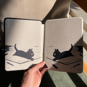 Puede incluir: Funda para lector de libros electr&oacute;nicos negra y beige con una ilustraci&oacute;n de un gato negro en cada lado. El gato est&aacute; tumbado sobre un libro con un bol&iacute;grafo, libros y una taza de caf&eacute;. La funda est&aacute; abierta, revelando el interior.