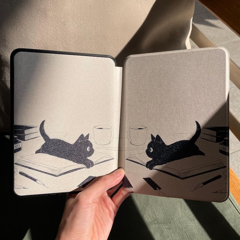 Puede incluir: Funda para lector de libros electr&oacute;nicos negra y beige con una ilustraci&oacute;n de un gato negro en cada lado. El gato est&aacute; tumbado sobre un libro con un bol&iacute;grafo, libros y una taza de caf&eacute;. La funda est&aacute; abierta, revelando el interior.