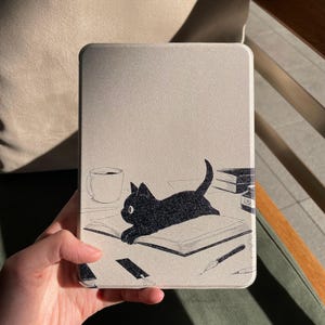 Puede incluir: Una funda de tableta de color claro con una ilustración de un gato negro tumbado en un libro abierto. El diseño incluye una taza de café, un tintero y un bolígrafo. La funda está sujeta por una mano.