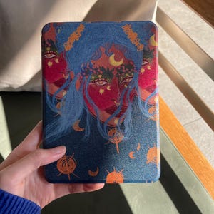 Könnte beinhalten: Eine blaue und rote dekorative Tablet-Hülle mit einer himmlischen Illustration. Das Kunstwerk zeigt drei Gesichter mit blauem Haar, Monden und sonnenartigen Symbolen. Die Hülle hat eine strukturierte Oberfläche.