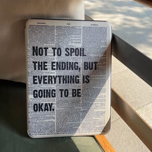 Könnte beinhalten: Eine rechteckige Tablet-Hülle mit Zeitungsmuster. Der Text "NOT TO SPOIL THE ENDING, BUT EVERYTHING IS GOING TO BE OKAY" ist in fetten schwarzen Buchstaben auf der Vorderseite gedruckt.