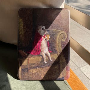 Peut inclure: Une coque rectangulaire avec une illustration fantaisiste d'une créature ressemblant à un chien, enveloppée dans une couverture rouge, tenant une tasse jaune. L'œuvre est située dans un intérieur de style vintage avec une chaise à motifs.