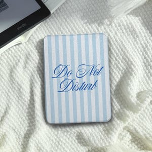 Può includere: Una custodia rettangolare per e-reader con un motivo a righe verticali azzurre e bianche. Il testo "Do Not Disturb" è stampato in un carattere corsivo blu scuro al centro della copertina, che presenta una finitura liscia e opaca.