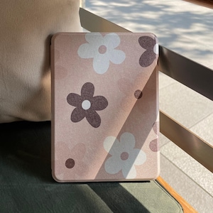 Könnte beinhalten: Eine rechteckige Hülle mit floralem Muster in einem sanften Rosaton. Die Hülle hat ein sich wiederholendes Design aus weißen und braunen Blumen mit kleinen braunen Punkten. Die Hülle liegt auf einer grünen Oberfläche.