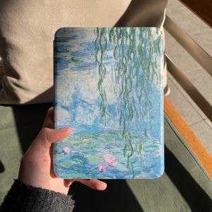Könnte beinhalten: Eine Tablet-Hülle mit einem Monet-Gemälde von Seerosen in Blau, Grün und Rosa. Das Kunstwerk zeigt eine Teichszene mit Seerosenblättern und Trauerweidenzweigen. Die Hülle wird von einer Person gehalten.