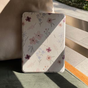 Könnte beinhalten: Tablet-Hülle mit floralem Muster. Der cremefarbene Hintergrund zeigt rosa, lila und blaue Blumen. Die rechteckige Hülle mit abgerundeten Ecken liegt auf einem grünen Kissen.