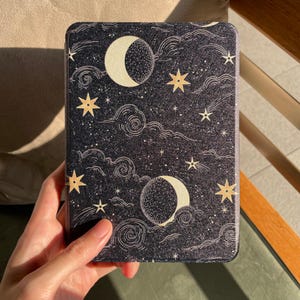 以下が含まれることがあります： 黒と白の電子書籍リーダーカバー。夜空に三日月、星、渦巻く雲が描かれたデザインです。カバーは手で持たれています。