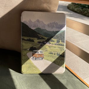 Könnte beinhalten: Eine rechteckige Hülle mit einer Landschaftsillustration von Bergen, einer Straße und einem orangefarbenen Oldtimer-Van. Die Hülle hat einen hellbeigen Hintergrund mit grünen und braunen Details und einen Schatten oben.