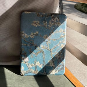 May include: A rectangular, blue case with a floral design inspired by Van Gogh's "Almond Blossoms" painting. The case has a soft, textured appearance with white and beige blossoms and branches against a blue background. The case is resting on a green surface.
