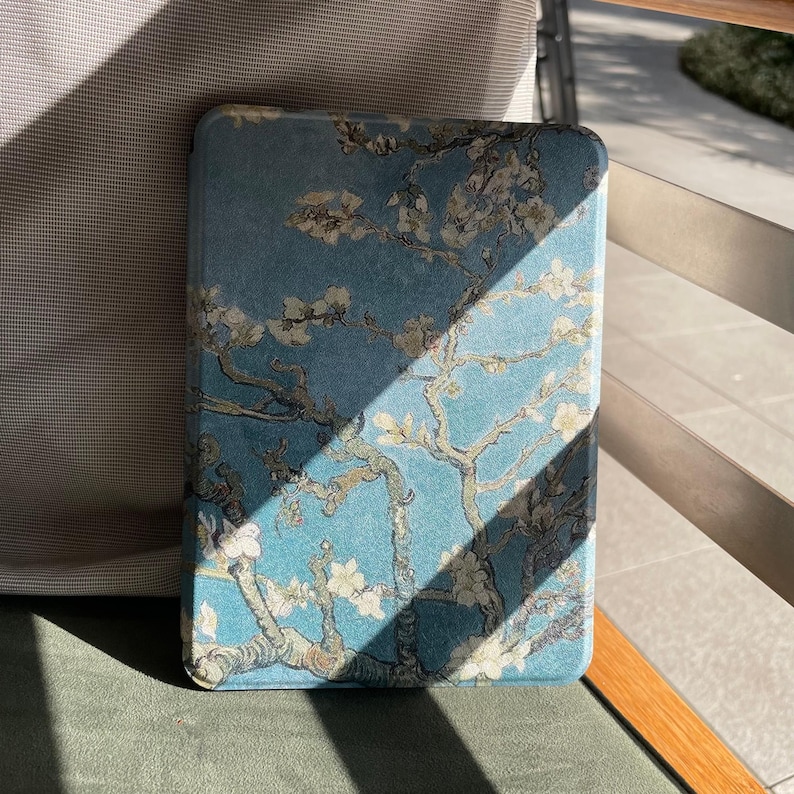 May include: A rectangular, blue case with a floral design inspired by Van Gogh's "Almond Blossoms" painting. The case has a soft, textured appearance with white and beige blossoms and branches against a blue background. The case is resting on a green surface.