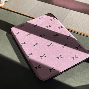 Op de afbeelding: Een roze tablethoesje met een patroon van zwarte strikvormen. De rechthoekige hoes met afgeronde hoeken ligt op een donkergroen oppervlak. De hoes wordt door zonlicht verlicht.