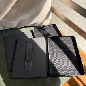 May include: Several grey tablet cases and trays are displayed. The cases have a smooth, matte finish and an elastic hand strap. The trays feature a textured, honeycomb pattern inside. The items are arranged on a green surface.