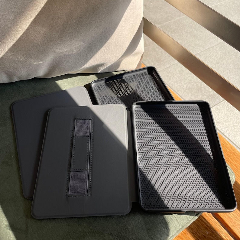 May include: Several grey tablet cases and trays are displayed. The cases have a smooth, matte finish and an elastic hand strap. The trays feature a textured, honeycomb pattern inside. The items are arranged on a green surface.