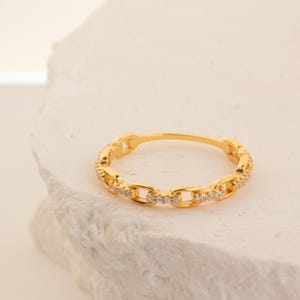 May include: A gold ring with a chain link design. The ring has small, clear stones set along the chain links. The ring is displayed on a white, textured surface.