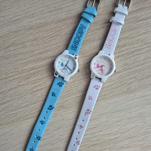 Puede incluir: Dos relojes de personaje Snoopy. Uno tiene una correa azul claro con huellas de patas y la palabra "SNOOPY" impresa. El otro tiene una correa blanca con huellas de patas rosas y la palabra "SNOOPY" impresa.