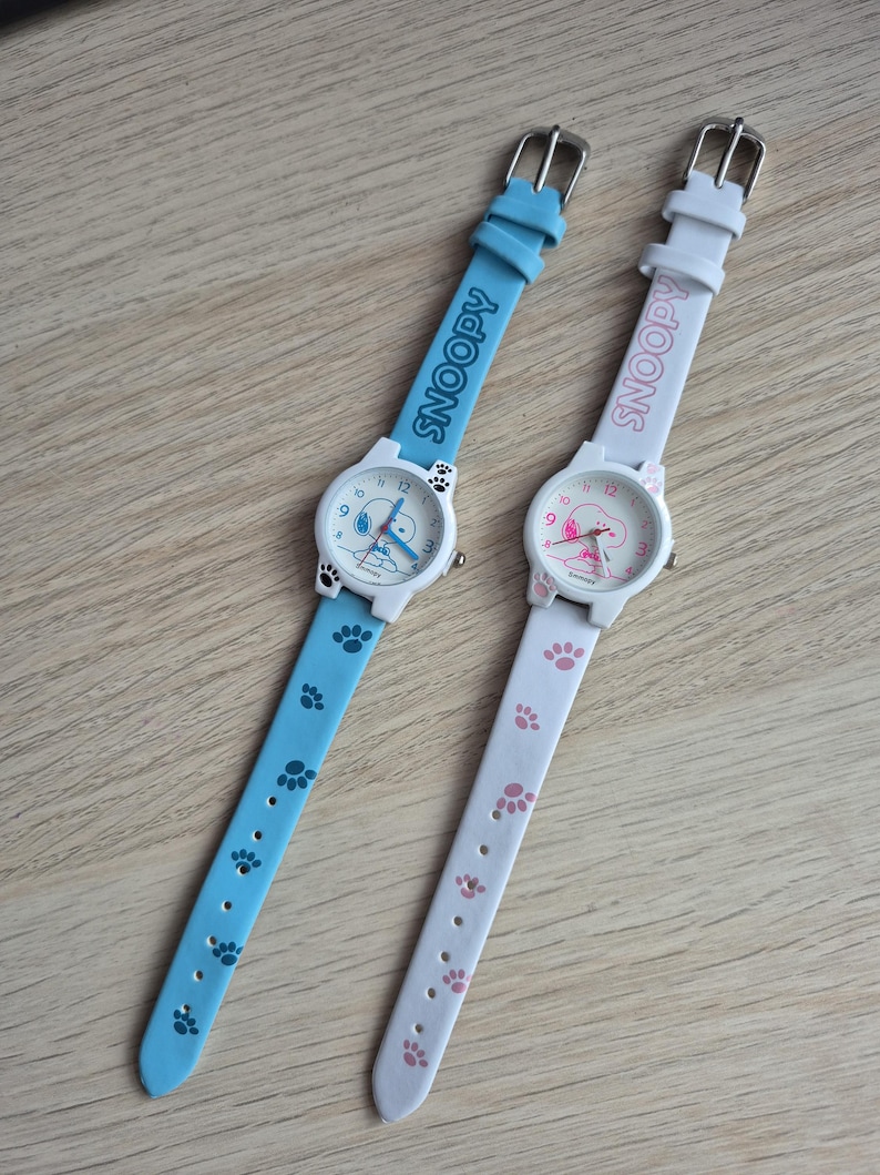 May include: Two Snoopy character watches. One watch has a light blue band with paw prints and the word "SNOOPY" printed on the band. The other watch has a white band with pink paw prints and the word "SNOOPY" printed on the band.