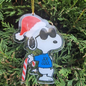 May include: A clear acrylic Christmas ornament featuring Snoopy wearing sunglasses and a Santa hat. Snoopy is wearing a blue shirt with the words "JOE COOL" and holding a candy cane. The ornament hangs from a gold string.