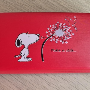 May include: A red glasses case with a Snoopy illustration blowing a dandelion, with the text "Make a wish...". The case has a rounded shape and a cartoon design.