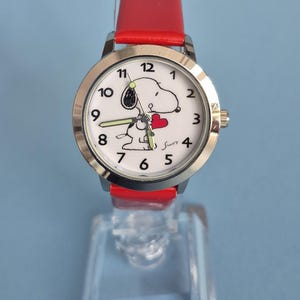 May include: A wristwatch with a bright red leather strap and a silver-tone metal case. The white dial features black Arabic numerals, a graphic of the character Snoopy holding a red heart, and luminous hands. The signature Schulz is printed in black.