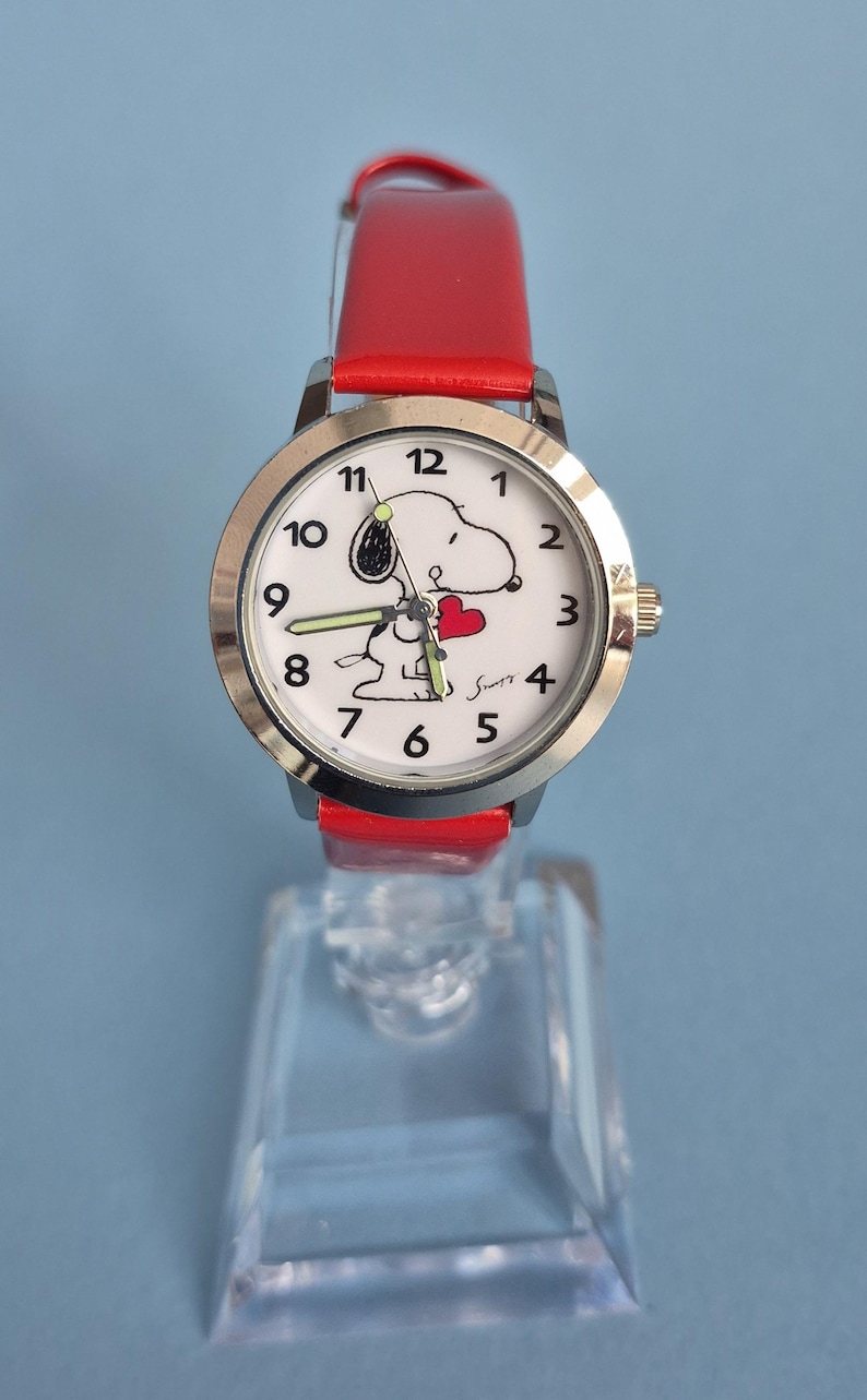 May include: A wristwatch with a bright red leather strap and a silver-tone metal case. The white dial features black Arabic numerals, a graphic of the character Snoopy holding a red heart, and luminous hands. The signature Schulz is printed in black.