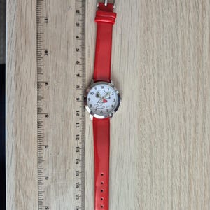 May include: A wristwatch with a bright red strap and a silver-tone case. The white watch face features a cartoon dog holding a red heart. A clear plastic ruler is placed alongside the watch, showing a total length of approximately 8.5 inches.